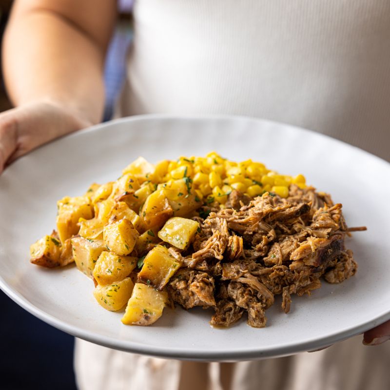 Close-up shot of Pulled Pork with Potato Salad being held up on a fork by a woman wearing a white shirt and cream-colored shorts. The tender pulled pork and creamy potato salad are in focus, showcasing their texture and vibrant colors.