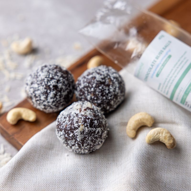 Three chocolate protein balls covered in dessicated coconut, sitting on a light tan cloth and wooden board, surrounded by cashews, a sprinkle of dessicated coconut and the wrapping label the protein balls are delivered in.