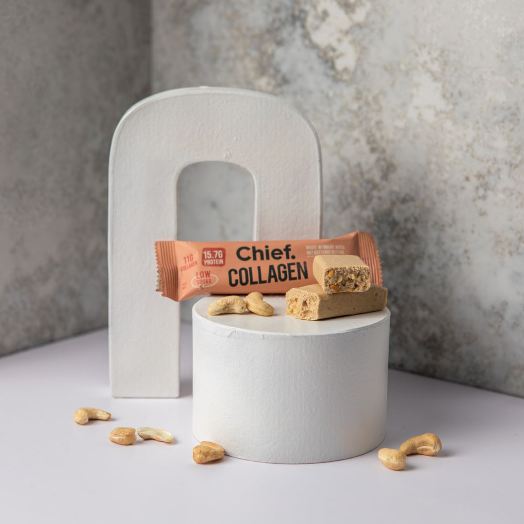 Against a marble backdrop, a cream coloured cashew bar, broken in half, sits atop a cylindrical prop with it's packaging placed behind it and cashews scattered around.