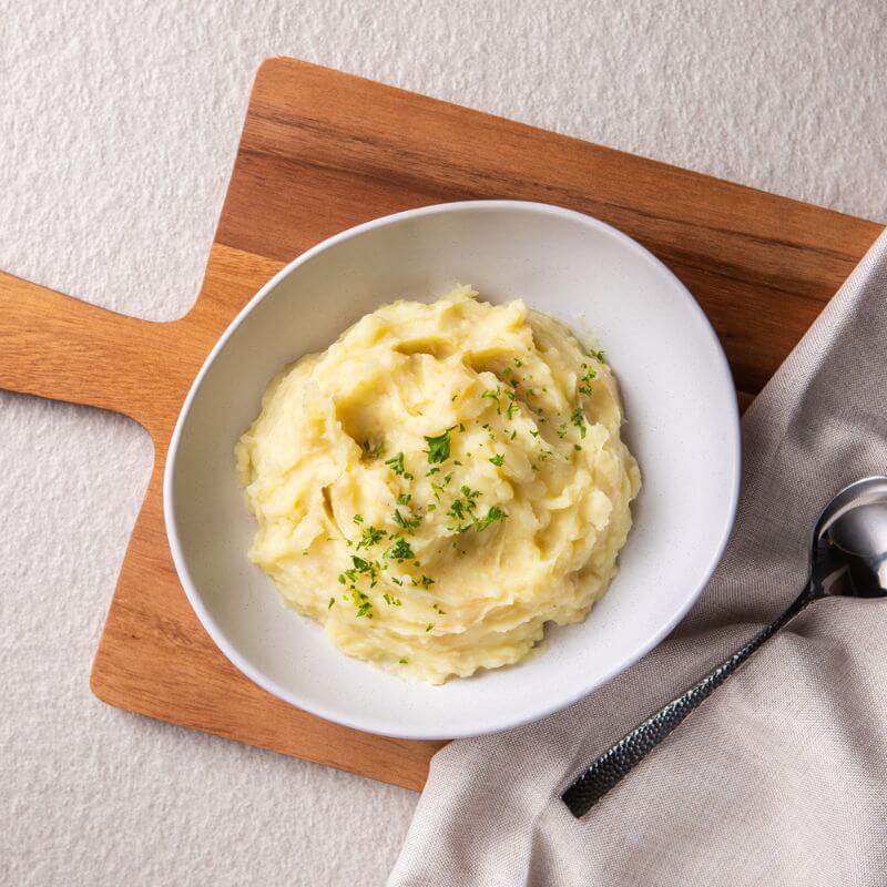 A white bowl filled with creamy mashed potato sits atop a grey brown cloth on a wooden board with a serving spoon to the right, all placed on a white table.