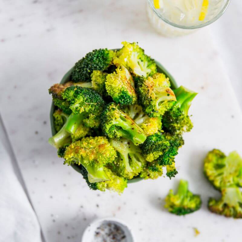 A top-down view of lightly charred broccoli overflowing a bowl served on a marble bench top.