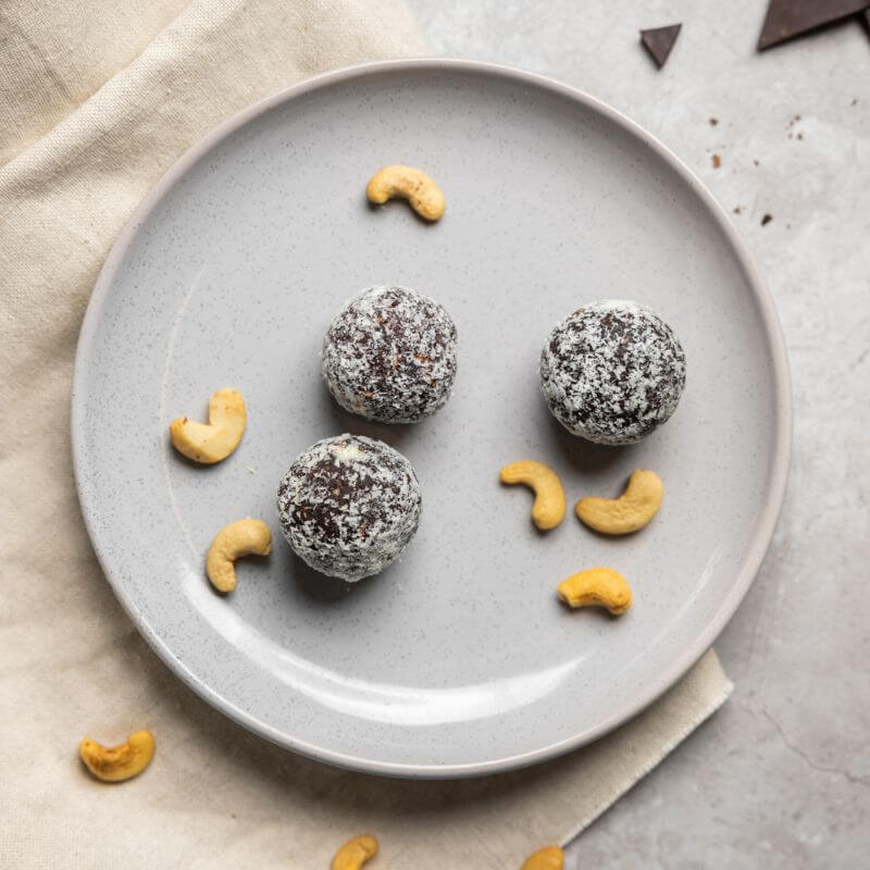 A top-down photograph of three chocolate protein balls covered in dessicated coconut and surrounded by cashews on a white plate, placed on a tan coloured cloth on a marble bench top.