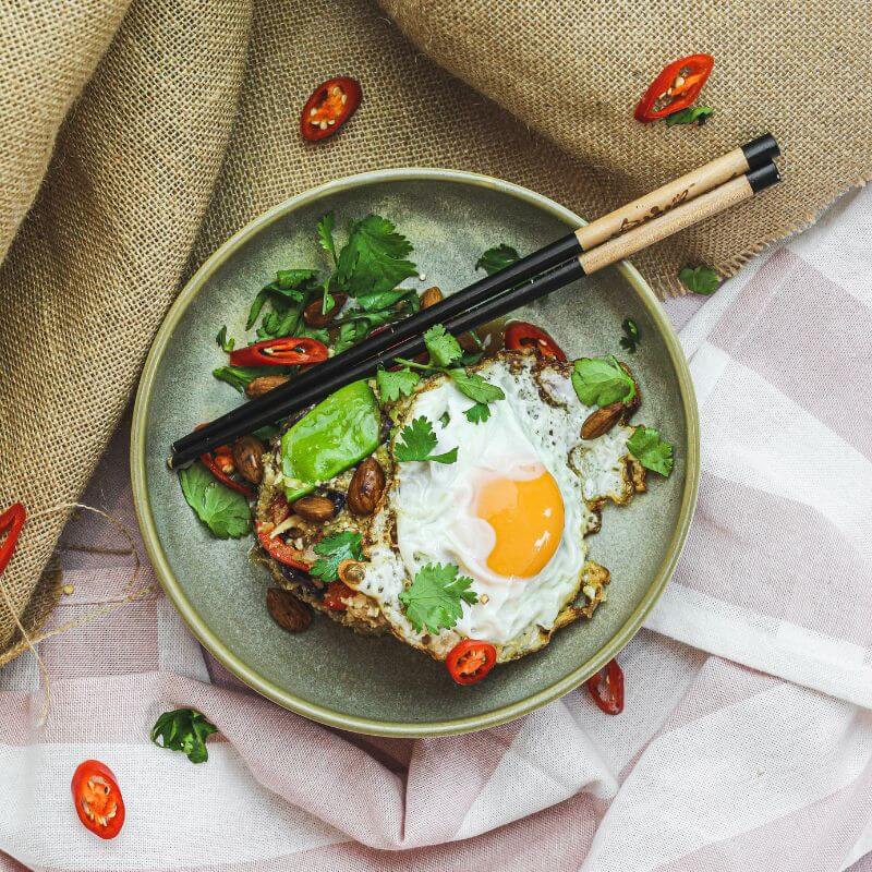 A top down photograph of our vegetarian Nasi Goreng, topped with a fried egg and sprinkled with fresh coriander and chilli, all served in a green bowl with chopsticks. The bowl is placed on a striped white cloth with a matching hessian cloth beside, and some extra chilli.