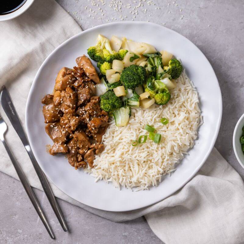 Flat lay shot of Teriyaki Chicken on a white plate with a beige cloth underneath, set on a grey marble countertop. The dish features tender stir-fried chicken thigh in a sticky, sweet teriyaki sauce, served on basmati rice with steamed broccoli and bok choy. Sesame seeds are scattered on top as garnish. A fork is placed to the left of the plate.