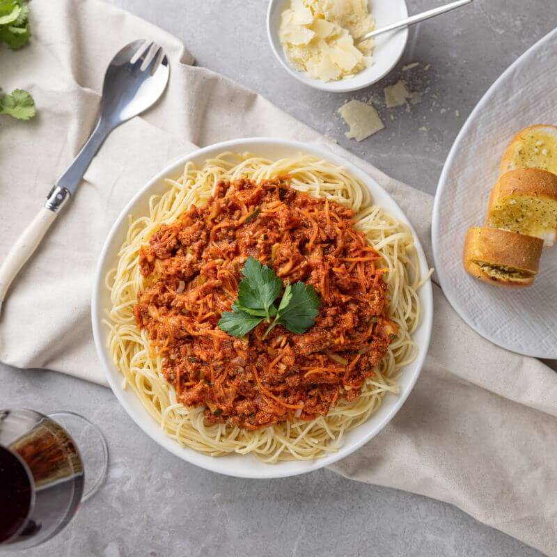 Flat lay shot of Spaghetti Bolognese on a white plate, garnished with parsley, surrounded by garlic bread on a side plate, a ramekin of fresh Parmesan cheese, a serving spoon, and a glass of red wine.