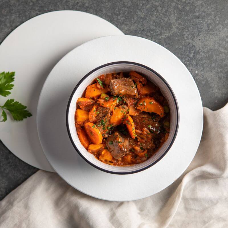 A top-down view of Slow Cooked Beef Stew in a white bowl with a black rim, placed on a white saucer plate. A cream-colored cloth is tucked underneath, and the bowl rests on a dark grey marble counter. The stew is garnished with chopped spring onion, with fresh parsley leaves scattered in the background as a decorative touch.