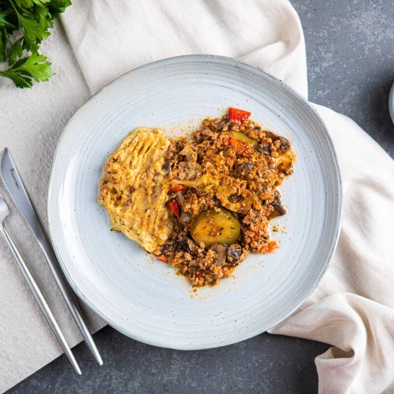 Flat lay shot of Savoury Mince with Baba Ganoush served on a blue-hued plate. The dish features fried mince, mushrooms, capsicum, tomato, onion, and herbs, topped with a creamy dollop of homemade baba ganoush. The plate rests on a beige cloth atop a dark marble countertop, with a fork and knife placed to the left. Parsley is scattered in the background, adding a touch of freshness