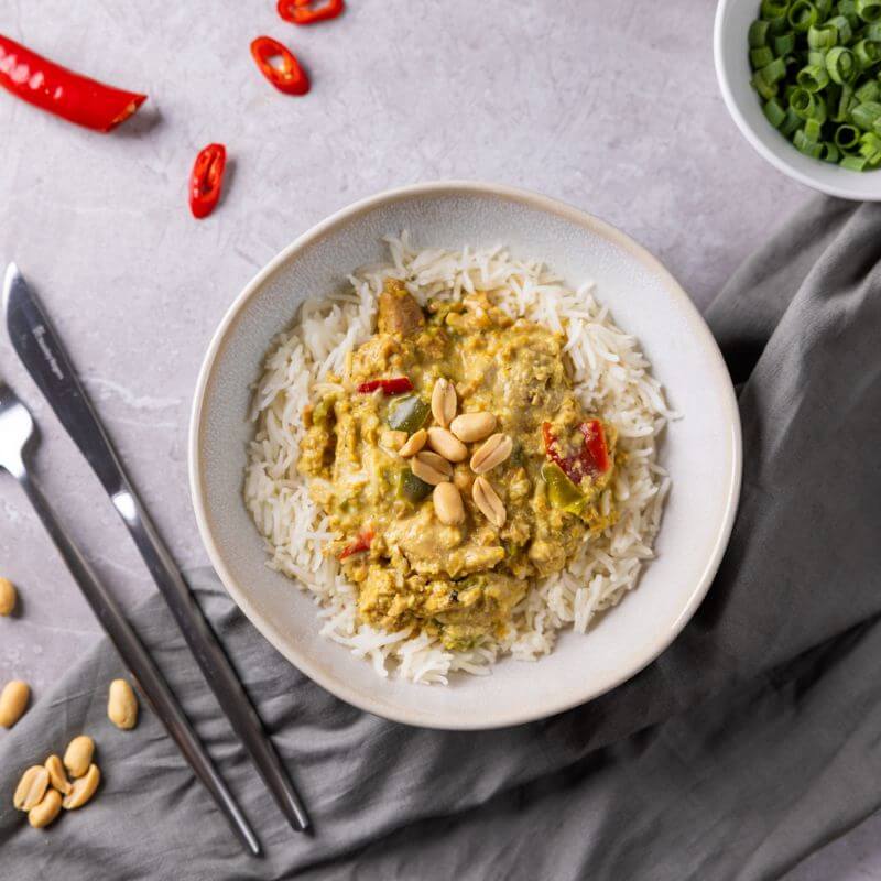 A top down photograph depicting a cream coloured ceramic bowl containing basmati rice and a rich, creamy satay sauce with chicken, capsicum and peanuts. The bowl is placed on a grey marble bench top with a green grey cloth beneath it, and is surrounded by a knife and fork, chopped chilli, and a white ramekin of chopped spring onions.