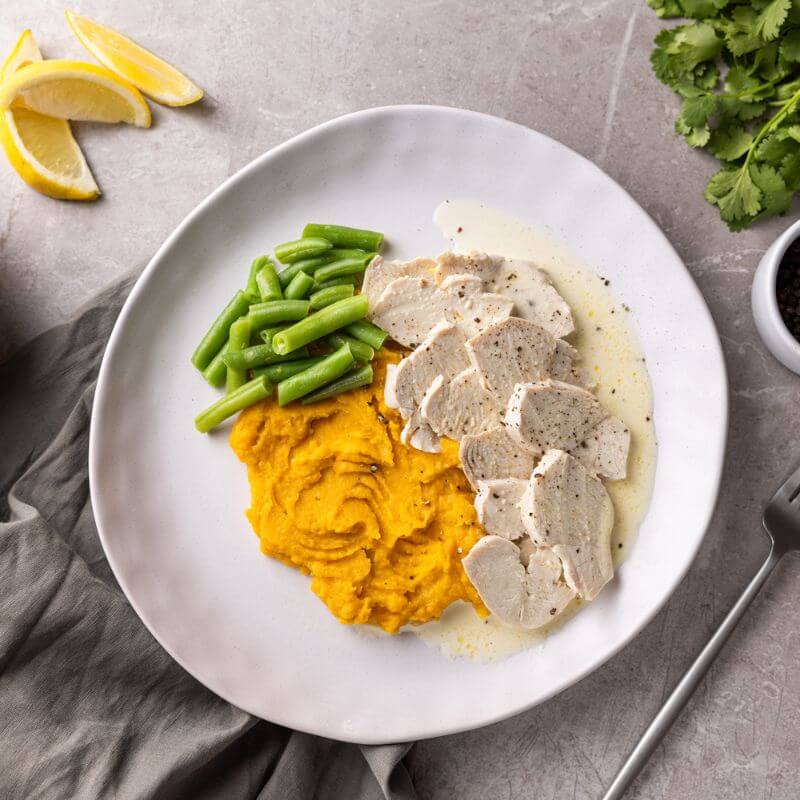 A top-down view of Lemon Pepper Chicken served on a white plate, sitting on a grey marble countertop with a green cloth tucked underneath. The dish features succulent chicken breast, green beans, and creamy lemon pepper sauce on top of sweet potato mash. A fork is placed to the right of the plate, with sliced lemon wedges on the counter and a white ramekin of cracked peppercorn nearby. A bunch of parsley is visible in the corner.