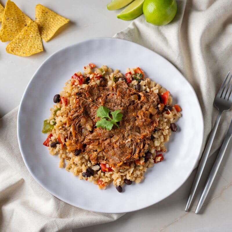 A top-down view of Chipotle Shredded Beef served on a white plate. The dish features tender pulled beef in a smoky adobo sauce served over Mexican-style cauliflower rice, garnished with a cilantro leaf. Corn chips are scattered around the plate, with sliced and whole lime wedges adding a vibrant touch. A fork and knife rest to the right of the plate, which is set on a beige cloth and a white benchtop.