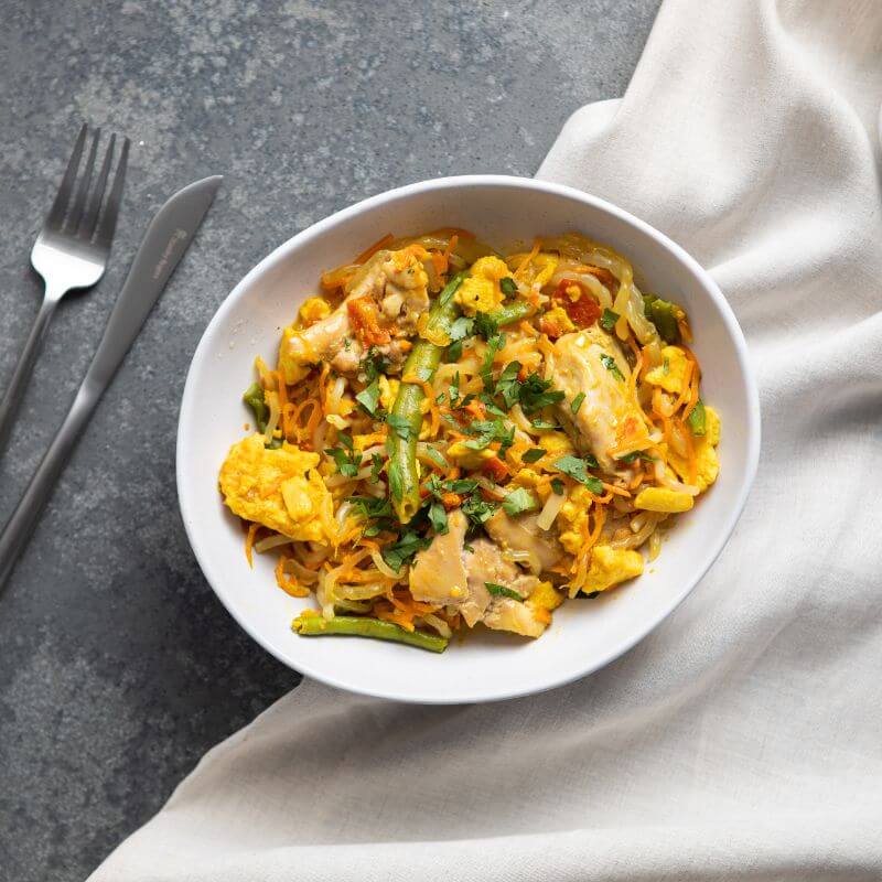 A top-down view of Chicken Singapore Noodles served in a white bowl on a dark grey benchtop. A white cloth rests underneath the bowl. Chopped spring onion is sprinkled on top of the noodles as a garnish. A fork and knife are placed to the left of the bowl.