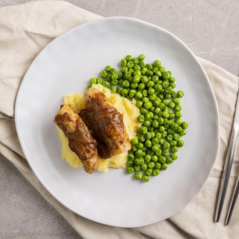 Bangers and Mash in a clear meal container, showcasing the sausages with onion gravy, mashed potatoes, and peas, placed on a neutral-colored placemat for a simple, appetizing presentation.