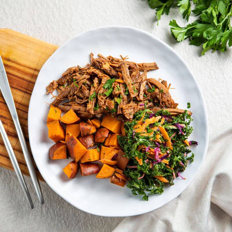 A top down photograph of pulled brisket in a barbeque sauce, roast sweet potato and a kale slaw on a white ceramic plate. The plate is resting on a chopping board and rough white bench top, with a knife and fork, a white cloth and green herbs on the side.