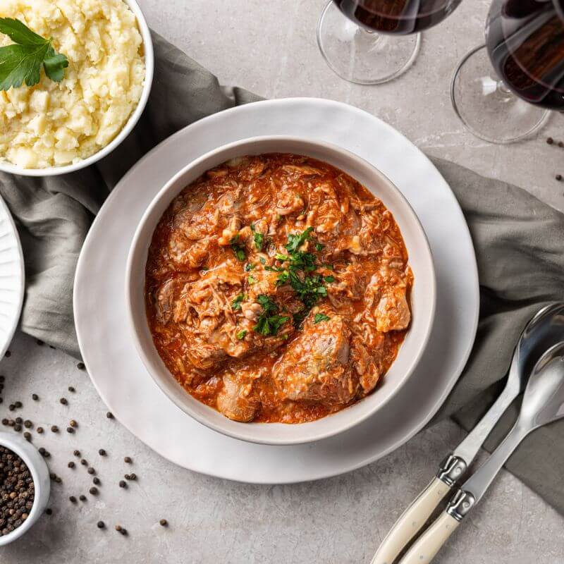 A top down photograph of lamb shanks off the bone in a rich tomato sauce, served in a white ceramic bowl on a white plate. Beside is a bowl of creamy white mashed potato, a silver fork and spoon, and all served upon a grey marbel bench with a darker grey cloth underneath.