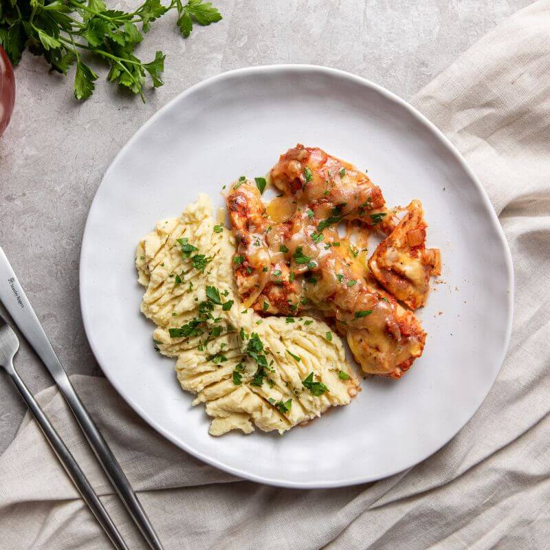 A top down photograph of a cheese and tomato sauce covered chicken breast, served with a creamy white mashed potato. A knife and fork lay to the side, and the dish is served on a marble bench with a cream cloth underneath.