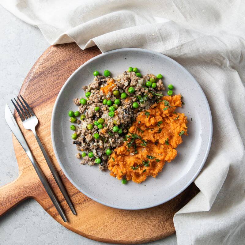 A top-down view of De-constructed Shepherd’s Pie served on a grey plate. The dish features a comforting blend of mashed potatoes, minced meat, and vegetables, garnished with chopped spring onions. The plate rests on a circular wooden cutting board with a beige cloth tucked underneath, placed on a grey marble benchtop. A fork and knife are positioned to the left of the plate.