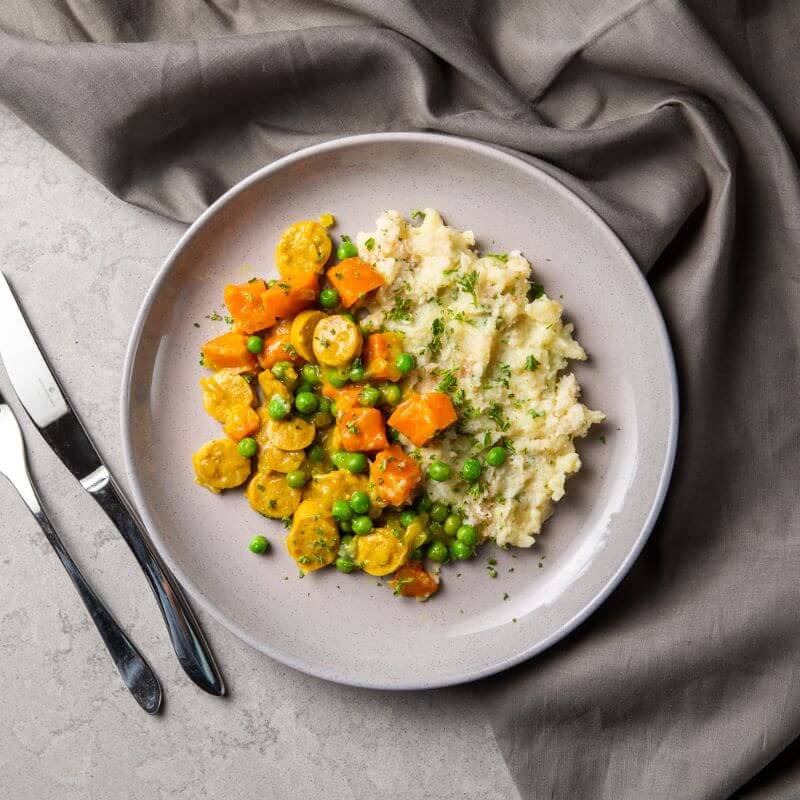 Flat lay image of curried sausage with cauliflower mash on a grey plate. The plate is against a dark grey cloth on a dark grey countertop. A fork and knife are placed to the left of the plate, while the curried sausage with cauliflower mash is garnished with chopped parsley on top for a fresh touch.