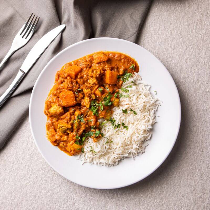 Flat lay image of Butter Chicken with Basmati Rice served on a clean white plate. The plate is set on a grey countertop with a dark grey cloth positioned to the left, adding contrast. A fork and knife rest on top of the cloth for a touch of elegance. The Butter Chicken is garnished with freshly chopped parsley, creating a pop of green against the creamy curry.