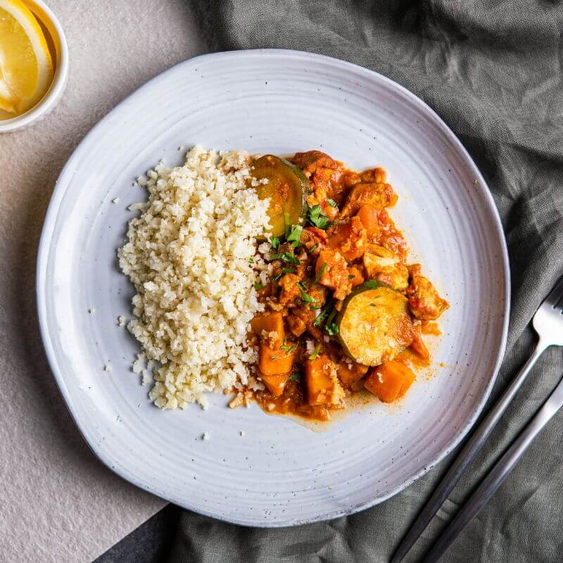 Flat lay image of Butter Chicken with Cauliflower Rice served on a clean white plate. The plate is set on a grey countertop with a dark grey cloth placed to the right, adding a cozy touch. A fork and knife rest on the cloth, and freshly chopped parsley is sprinkled on top of the butter chicken as a garnish. A white ramekin containing a lemon adds a fresh, zesty accent in the background.