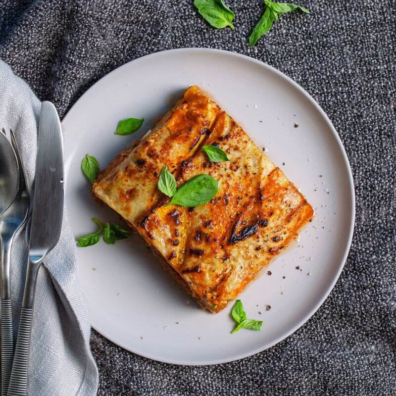 Flat lay of the Pumpkin Lasagna on a white plate, set against a dark blue cloth for a pop of color. A fork and knife are placed to the left of the plate, sitting on a light grey cloth. A few fresh spinach leaves are scattered on top of the lasagna as a garnish, adding a vibrant touch.