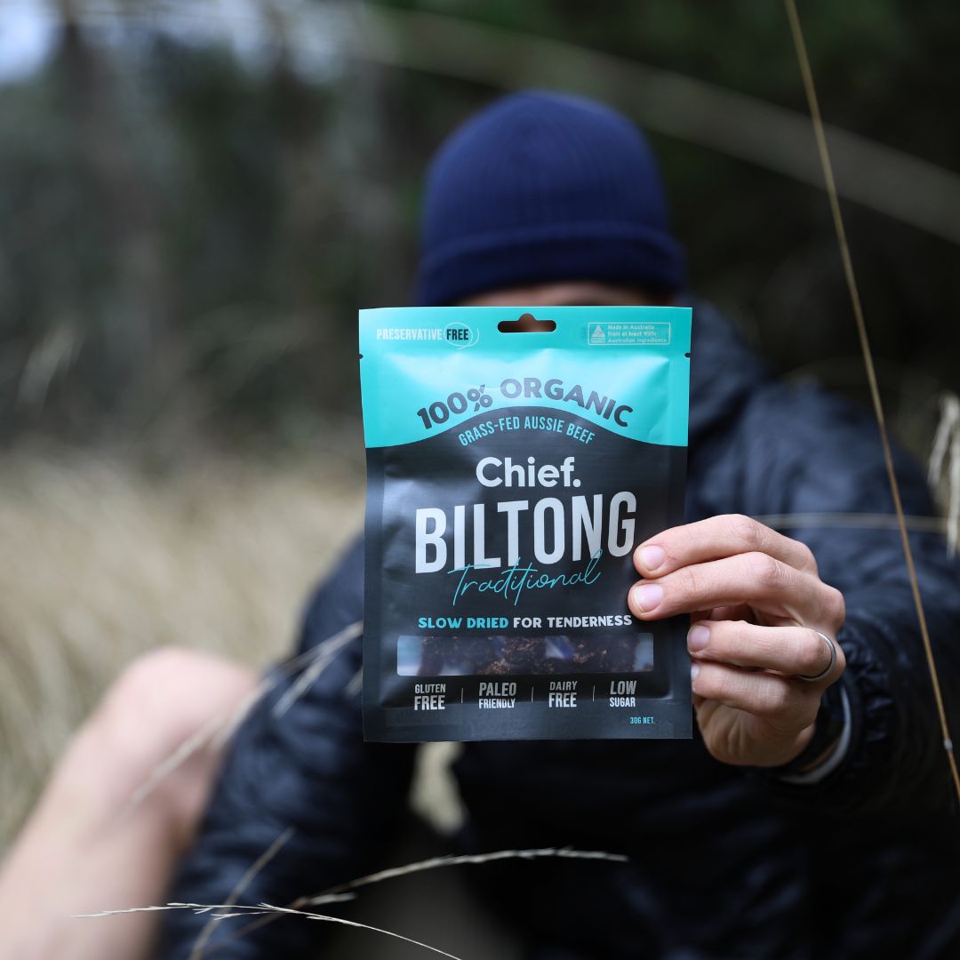 A close up of a packet of beef biltong being held by a person in a windproof jacket and a beanie, sitting in an outdoor field.