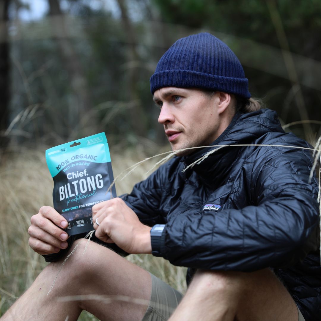 A young man in a waterproof windbreaker and beanie sitting in a field holding a packet of beef biltong.