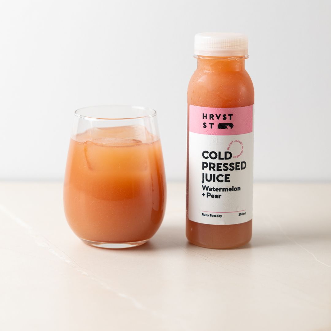 A drinking glass of a light pink juice, served beside a bottled serve of pink cold pressed juice, set against a white grey backdrop and bench.