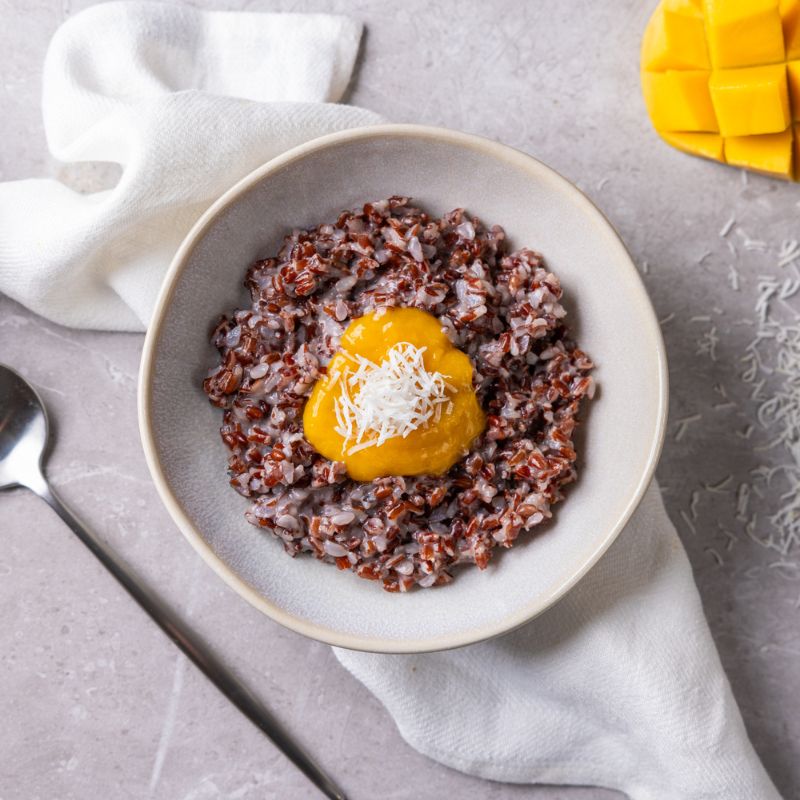 A top down photograph of a serve of red rice porridge, topped with mango puree and dessicated coconut in a plain, ceramic bowl. The bowl is sitting on a white cloth on a marble table, surrounded by sprinkles of dessicated coconut, a spoon, and a slice of fresh mango.