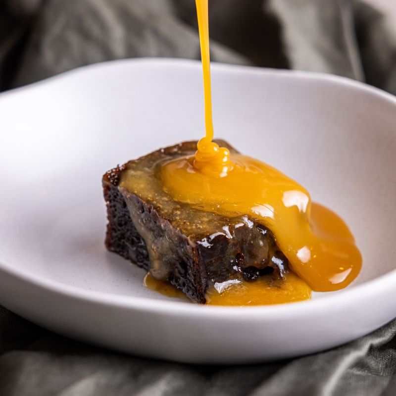 A single serve of rich sticky date pudding in a white ceramic bowl, being drizzled with a smooth, golden sticky date sauce.