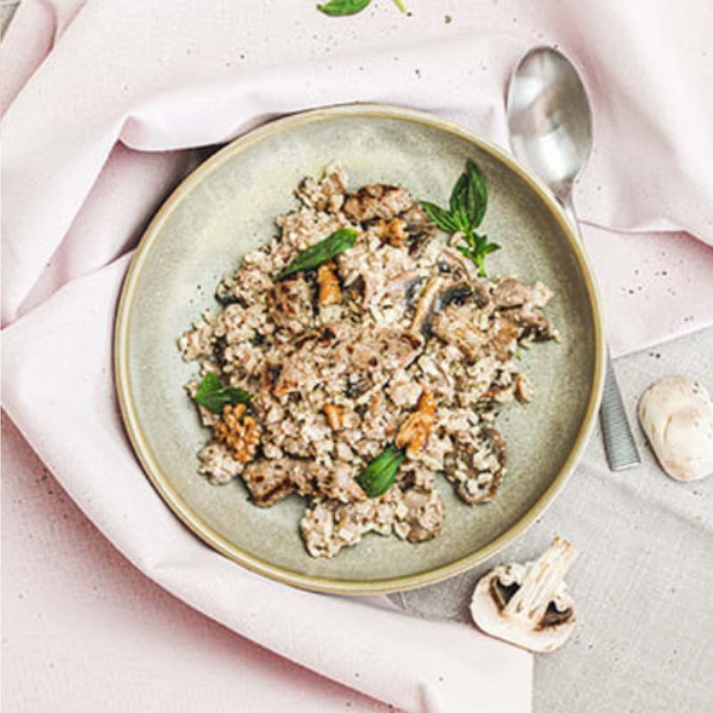 A top-down view of Chicken, Mushroom & Walnut Risotto served on a cream-colored plate. The creamy risotto, made with cauliflower rice, is topped with tender chicken, mushrooms, walnuts, and fresh spinach as a garnish. A silver spoon rests to the right of the plate, with a pink cloth tucked underneath. A sliced mushroom is visible in the background.