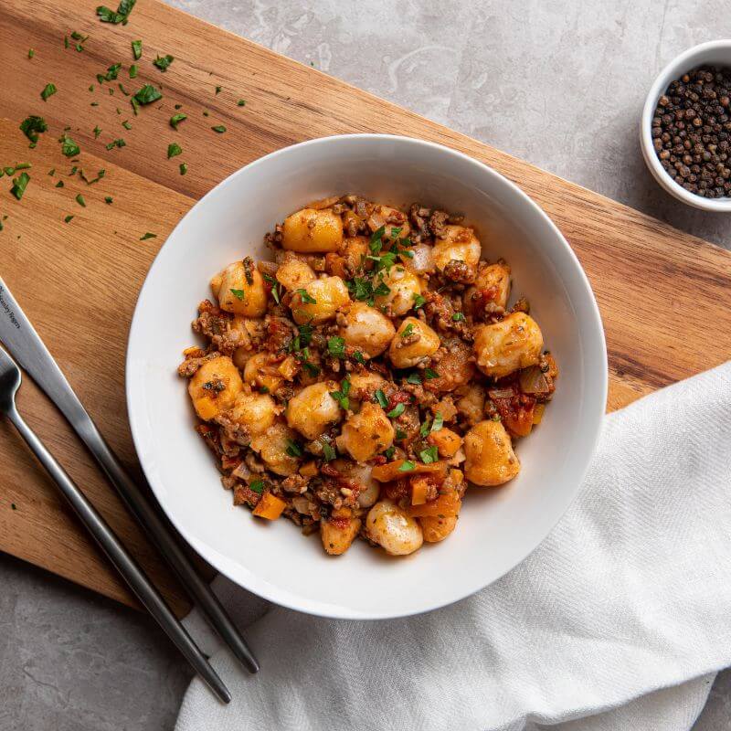 A top-down view of beef bolognese gnocchi served in a white bowl, placed on a wooden chopping board. A white cloth rests underneath the bowl, and chopped spring onion is sprinkled on the board and as a garnish on the dish. A white ramekin filled with black peppercorns sits in the background, and a fork and knife are placed to the left of the bowl.