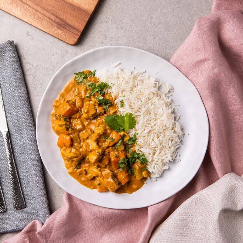 A top-down view of Chicken Masala with Basmati Rice served on a white plate. The creamy curry, featuring tender chicken coated in a spiced Masala gravy, is paired with fluffy basmati rice and garnished with chopped parsley. A fork and knife rest on a grey napkin to the left of the plate, with a pink cloth tucked underneath. The setup sits on a marble countertop, with a wooden chopping board faintly visible in the background.