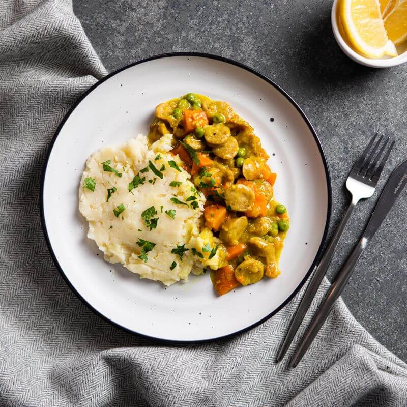 Flat lay image of curried sausage with white potato mash on a white/black-rimmed plate. The plate sits against a dark grey cloth on a dark grey countertop, providing a sleek contrast. A fork and knife are placed to the right of the plate, while a white ramekin with a sliced lemon and chopped parsley garnish are added for a touch of color.