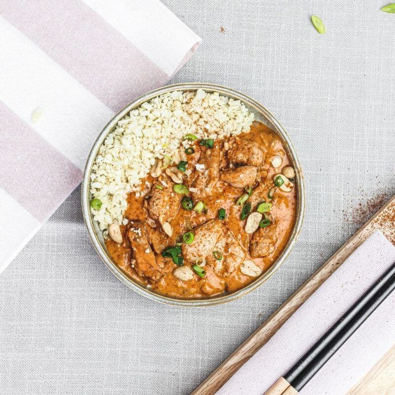 Flat lay image of Kung Pao Chicken served in a cream-colored bowl, resting on a light grey cloth. A beige and white striped cloth adds texture to the scene, and black chopsticks are placed to the right of the bowl. The dish is garnished with freshly chopped spring onion, adding a vibrant touch