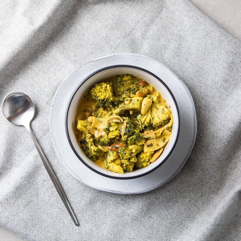 Flat lay shot of Thai Green Curry in a white bowl with a black rim, resting on a white plate. A spoon is placed to the left, all set on top of a grey cloth, showcasing the vibrant vegetables and creamy curry.