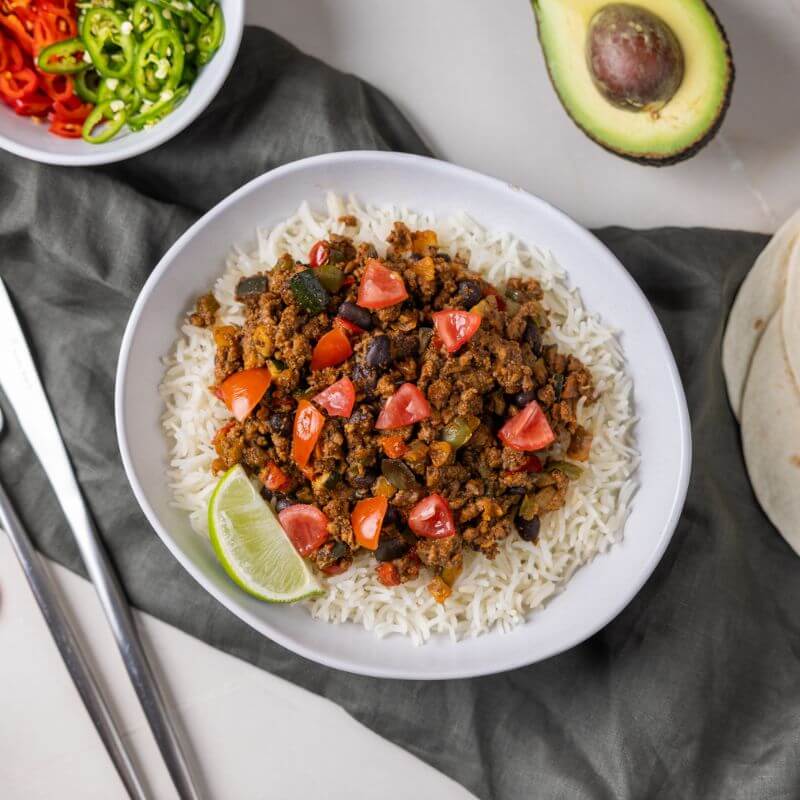 Flat lay image of the Burrito Bowl served in a clean white bowl. The bowl is placed on a dark grey cloth that sits atop a grey marble countertop, creating a stylish and cozy presentation. To the left of the bowl, a fork and knife are positioned neatly. A white ramekin filled with chopped red and green peppers, a sliced avocado, and tortillas are scattered in the frame to add authentic Mexican vibes and pops of vibrant color.