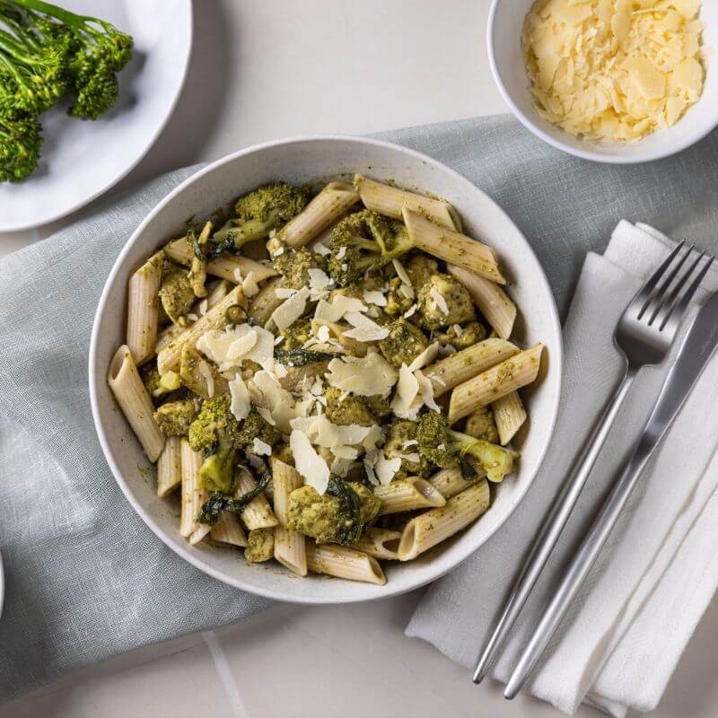 Flat lay shot of Chicken Pesto Penne in a white bowl, topped with freshly grated Parmesan cheese, accompanied by a fork and knife on a napkin, with a ramekin of Parmesan cheese and a plate of broccolini in the background.