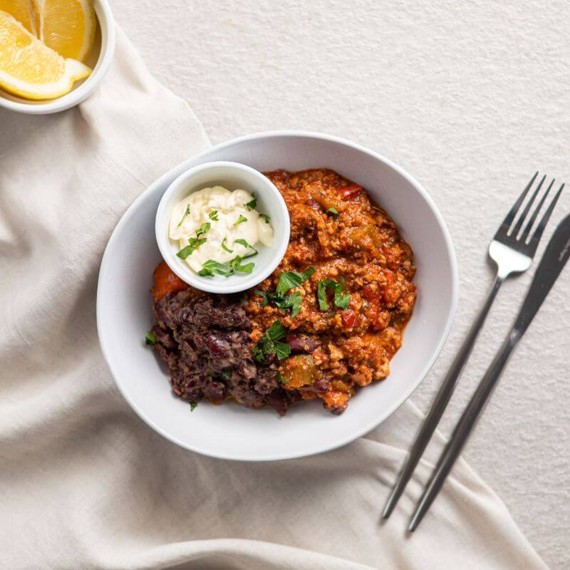 Flat lay shot of Chilli Con Carne in a white bowl, with a ramekin of sour cream topped with spring onion, and a garnish of spring onion on the chili. A knife and fork are placed to the right, and a ramekin of lemon is on the left.