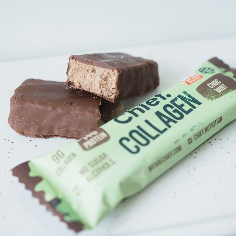 A side angled photograph featuring a green choc mint protein bar in the foreground with the bar itself cut in half placed behind. This is all set on a white marble tray.