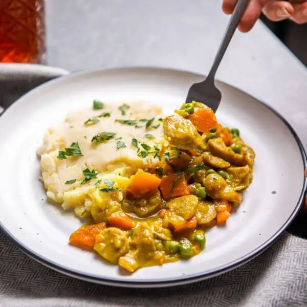 Action shot of curried sausage with white potato mash, with a fork digging into the sausage and mash, emphasizing the creamy texture and the hearty ingredients of the dish.