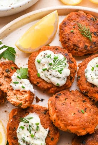 Cooked salmon cakes on a speckled platter, topped with garlic aioli, fresh herbs, and surrounded by cut lemon wedges.