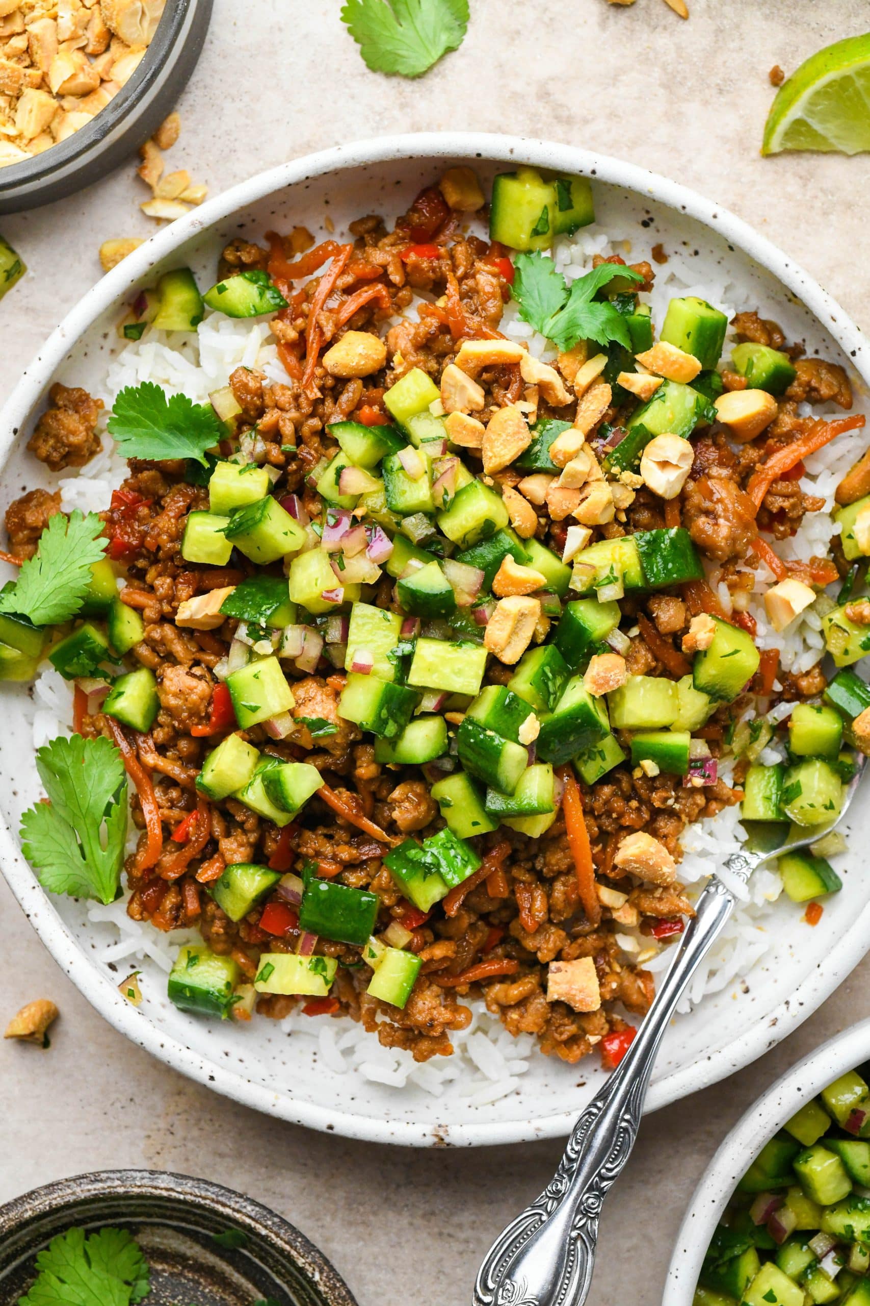 Asian ground pork and veggies in a bowl over white rice, with pickled cucumber salad, chopped cashews, and fresh cilantro.