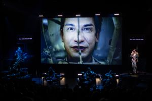 A multimedia performance setup with musicians in the foreground and a large screen showing a close-up of a man's face in an ominous setting, accompanied by superimposed subtitles like "Attach electric drills to the teeth!"