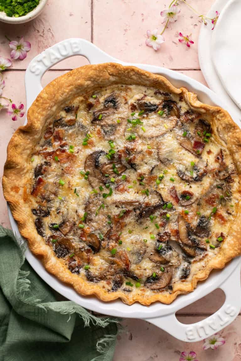 Bacon and mushroom quiche on a table with flowers and a napkin.