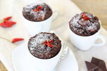 The image shows three chocolate mug cakes dusted with powdered sugar and garnished with small pieces of red chili peppers on top. The mug cakes are served in white coffee cups or mugs and appear to be freshly baked, with a rich, dark chocolate color. In the corner of the image, there are some pieces of chocolate bars and scattered red chili peppers, suggesting this is likely a spicy chocolate dessert that combines sweet and heat flavors. The cakes have a moist, cake-like texture and appear to have risen nicely in their mugs.