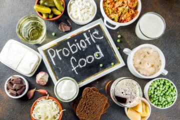 This image showcases a variety of probiotic foods arranged around a small chalkboard that says "Probiotic food." The assortment includes: Pickles in a small dish A bowl of cottage cheese A jar of sauerkraut Kimchi Glasses of yogurt and kefir A plate of soft cheese Dark chocolate squares Rye bread slices A bowl of peas A head of garlic Parmesan cheese pieces The overall theme emphasizes fermented foods and other natural sources of probiotics.