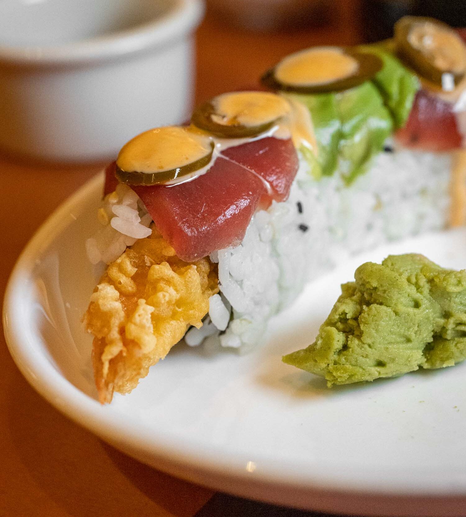 Close-up of a signature sushi roll composed of tempura shrimp and fresh tuna, garnished with thin sliced jalapeños, spicy mayo, and black sesame seeds, served with fresh wasabi and lettuce on a classic white ceramic plate in traditional Japanese style