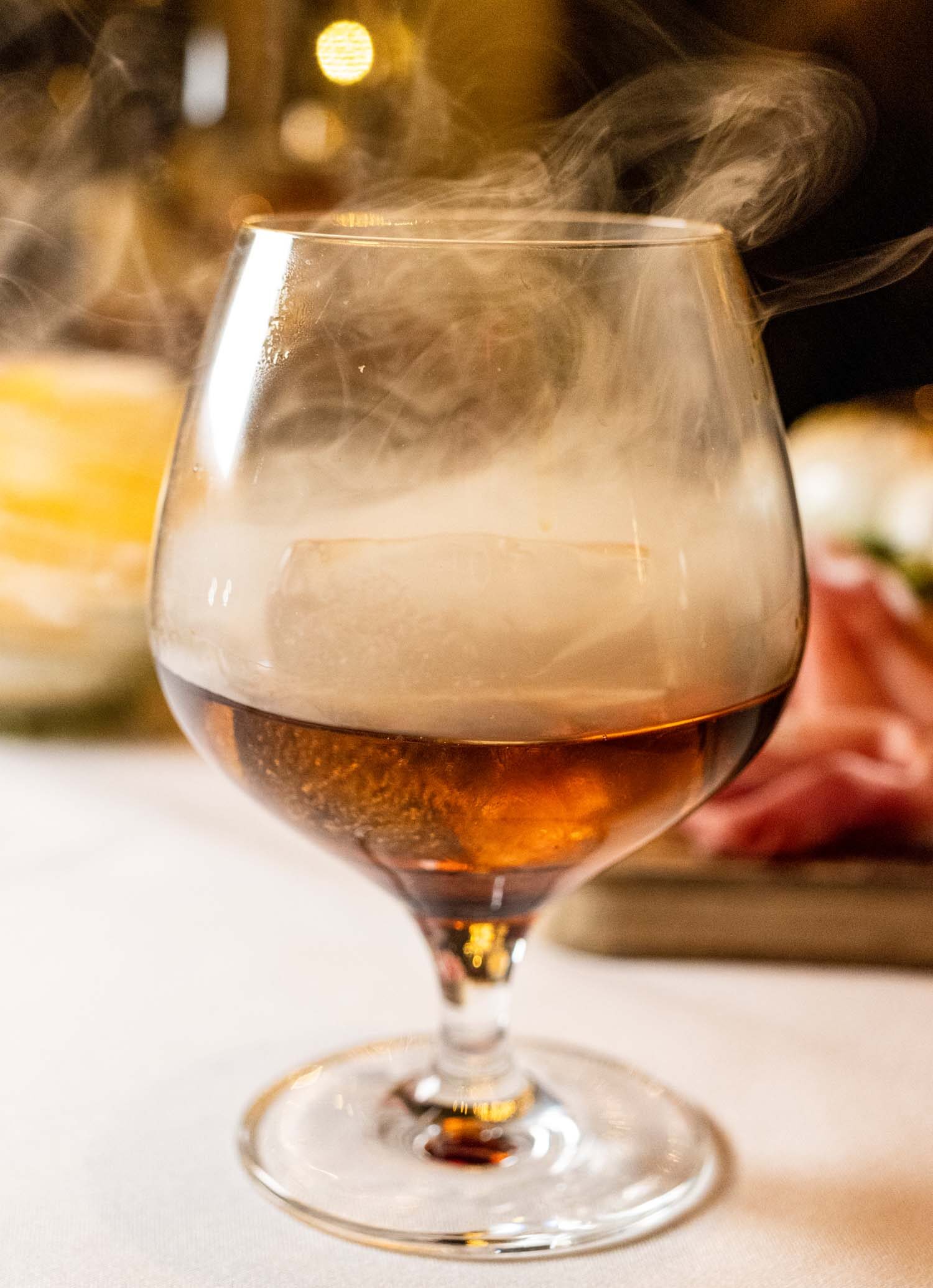 A smoky cocktail featuring Basil Hayden bourbon, Grand Marnier, and Luxardo cherry with walnut bitters, wisps of smoke swirling in a brandy snifter against a warm amber-lit background