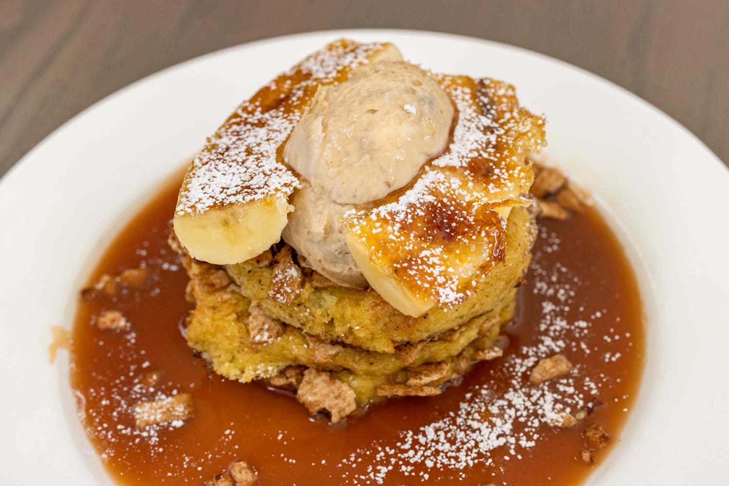 Stack of French toast topped with caramelized bananas, whipped cream, powdered sugar, and caramel sauce on a white plate