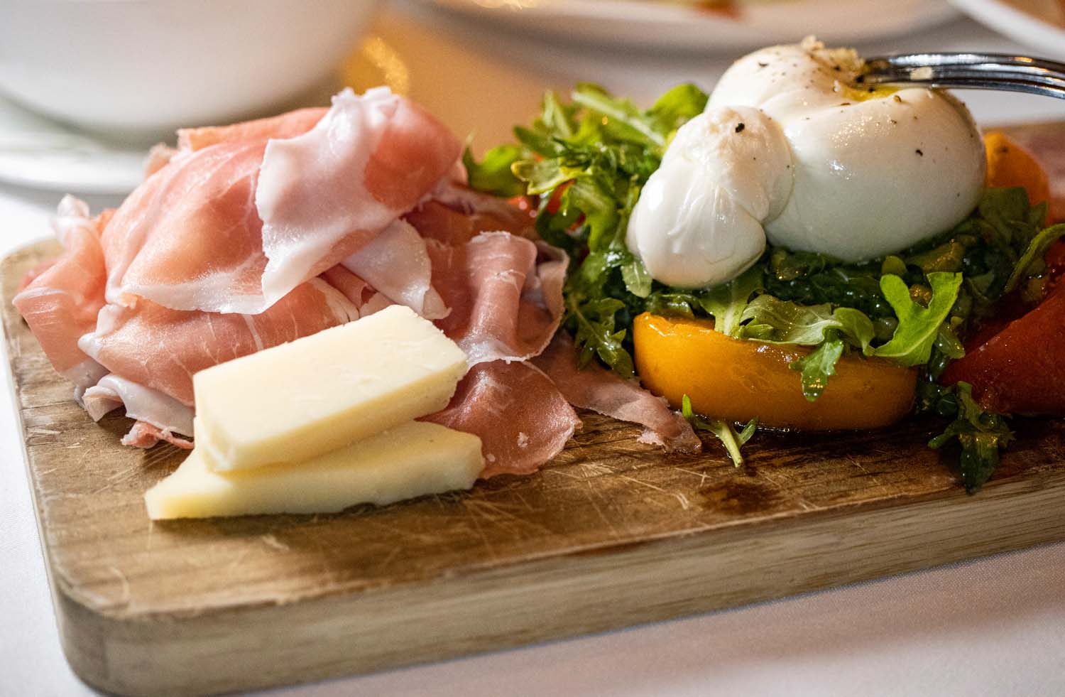 Rustic wooden board featuring thinly sliced prosciutto, fresh burrata cheese, arugula salad with heirloom tomatoes and fresh herbs, and two perfectly poached eggs, artfully presented on a weathered surface